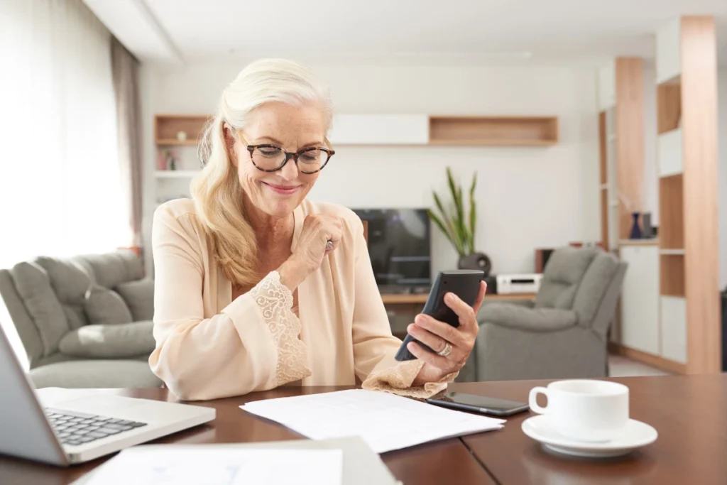 Smiling woman reviewing finances at home, using smartphone and paper budget tracker at a cozy kitchen table.