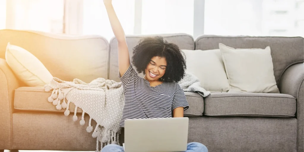 Woman smiling and raising fist in celebration while using a laptop, symbolizing the emotional boost from the debt snowball method.