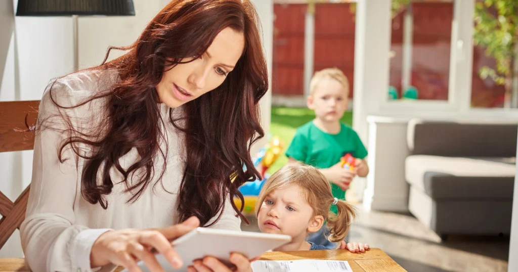 A tired mother calculating bills on a tablet with two young children in the background.
