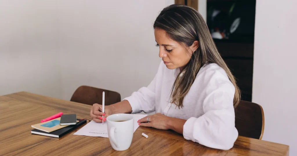 A woman sitting at a wooden table with a coffee cup and notebook, creating a zero-based budget.